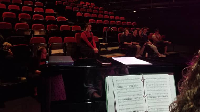 Students in theatre seats looking towards stage. Photo is taken from behind a person at a piano.
